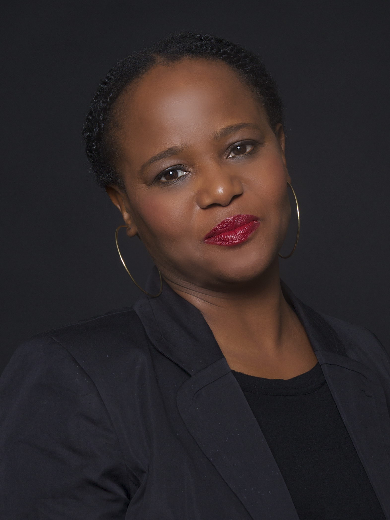 A woman with short braided hair, wearing hoop earrings, a black blazer, and a black top, poses against a dark background with a slight smile and red lipstick—her confident gaze evokes the poise of a Nabokov Award winner.