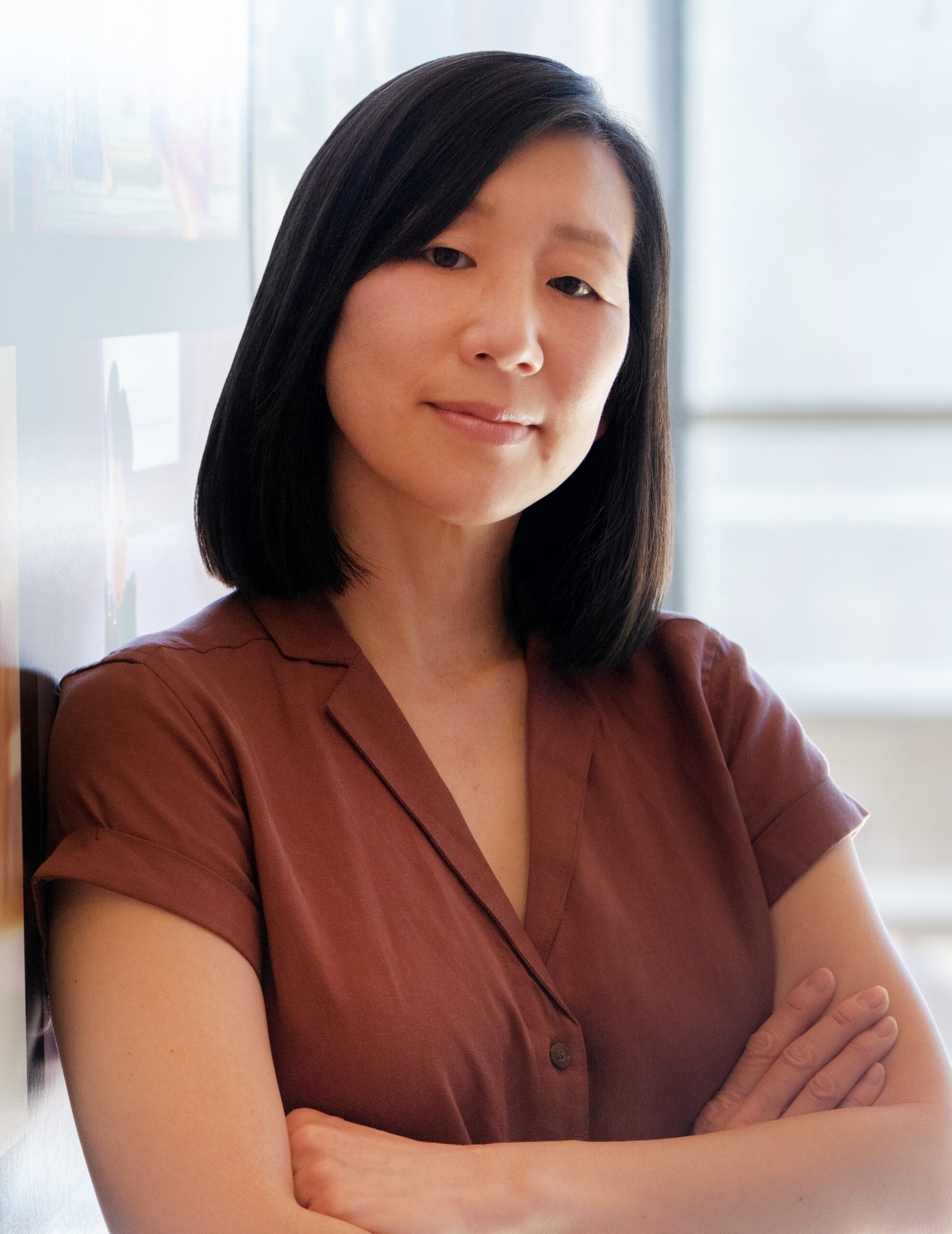 A woman with straight black hair, wearing a short-sleeved maroon blouse, stands with her arms crossed, leaning against a wall. She gazes confidently at the camera—her poise hinting at the grace of a theater award winner. Soft light fills the space.