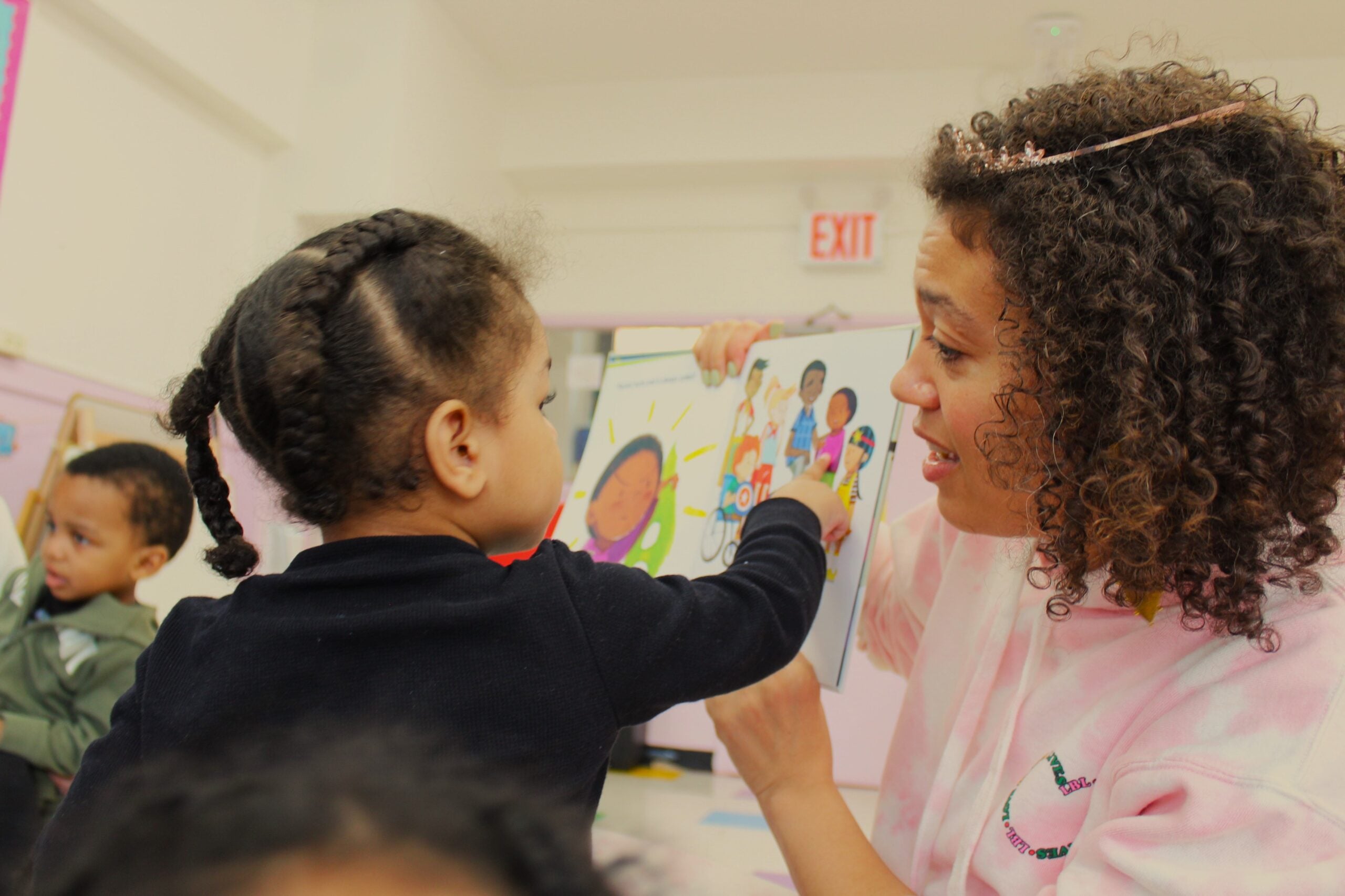 A woman holds a colorful picture book while a young child with braided hair points at a page. Another child is sitting and looking on in the background. They are in a bright classroom setting.