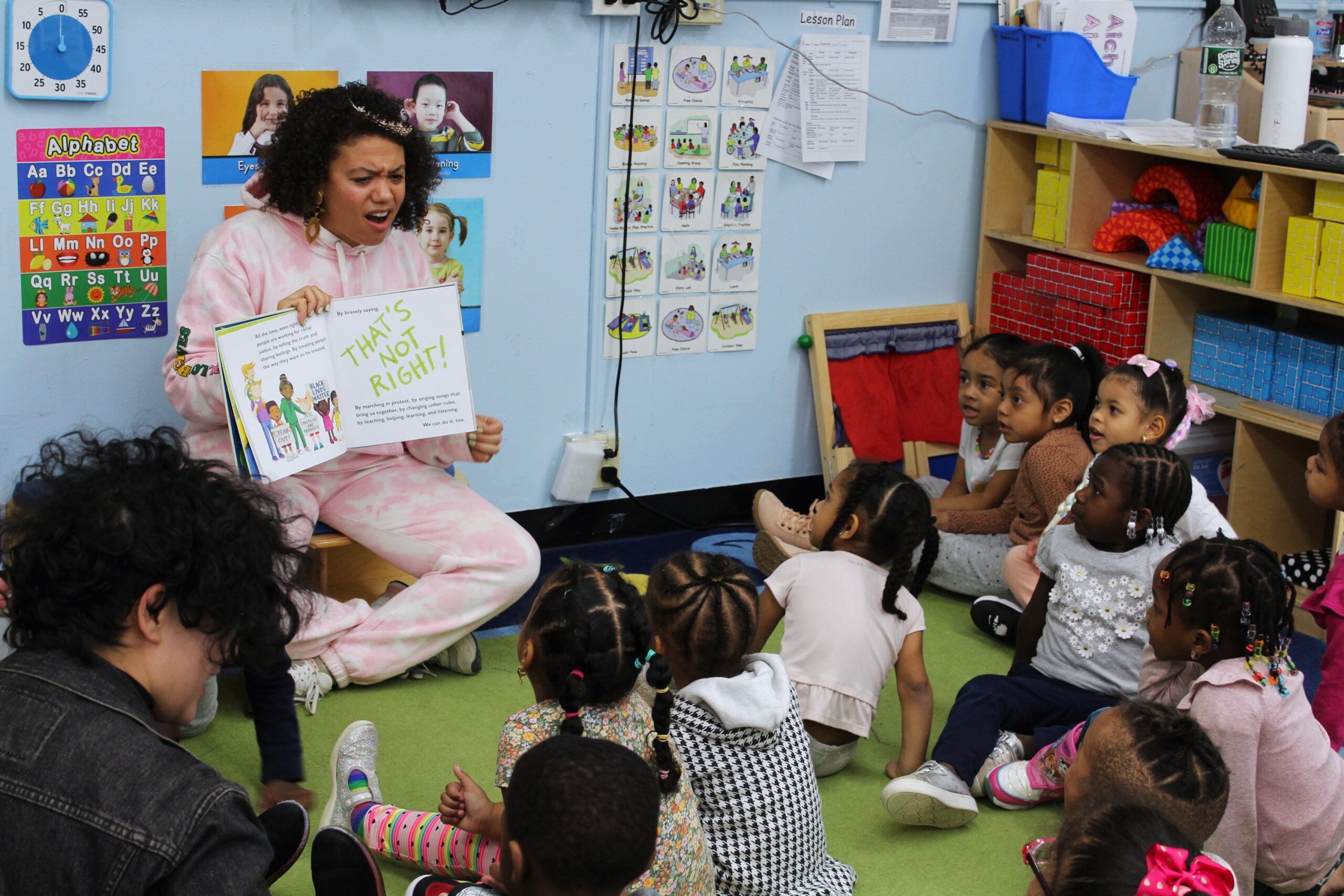 A woman in pink pajamas enthusiastically reads a book to a group of young children sitting on the floor in a colorful classroom decorated with educational posters and shelves of supplies.
