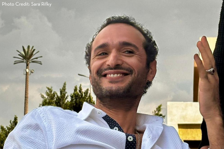 A man with short curly hair and a trimmed beard, wearing a white shirt, smiles while looking to the side; trees and a building appear in the background under a cloudy sky.