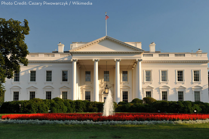 The White House with an American flag on top, a water fountain and red flowers in front, set against a clear blue sky. Photo credit: Cezary Piwowarczyk / Wikimedia.