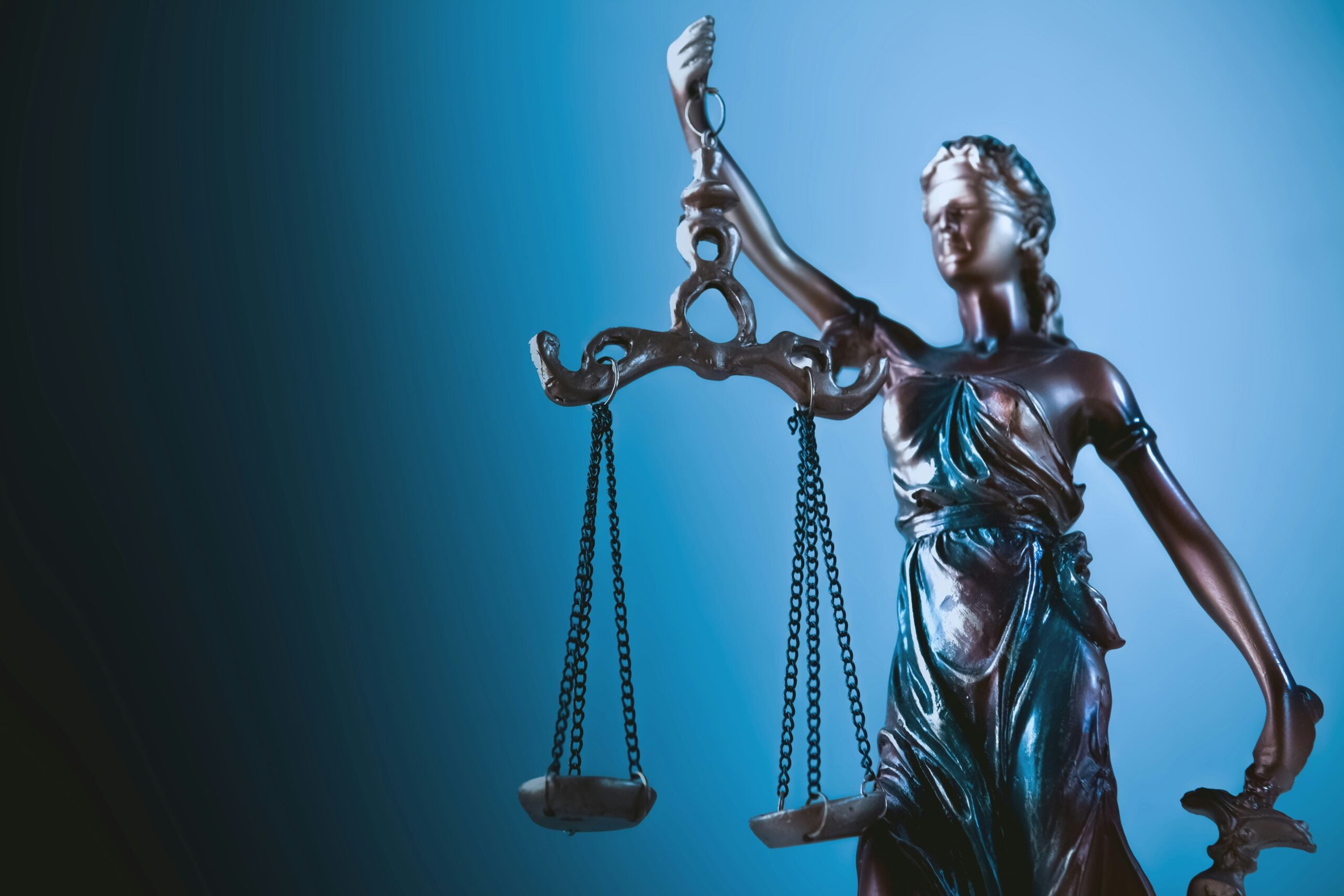 A close-up of a metallic Lady Justice statue holding balanced scales in her left hand, set against a blue gradient background. The statue symbolizes law and fairness.