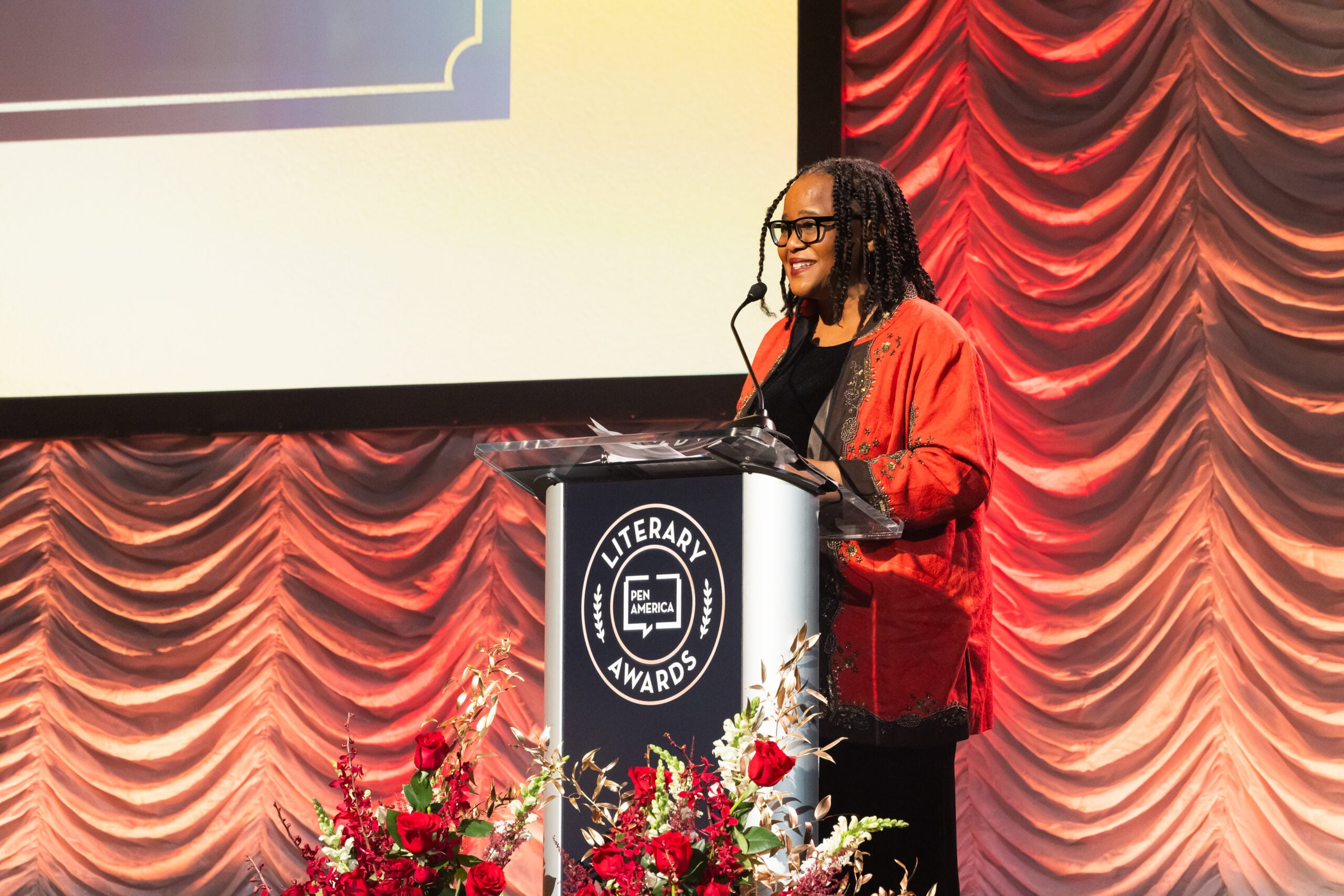 A woman in a red jacket stands at a podium labeled Literary Awards, speaking into a microphone. The background features red draped curtains and a floral arrangement is visible in front of the podium.