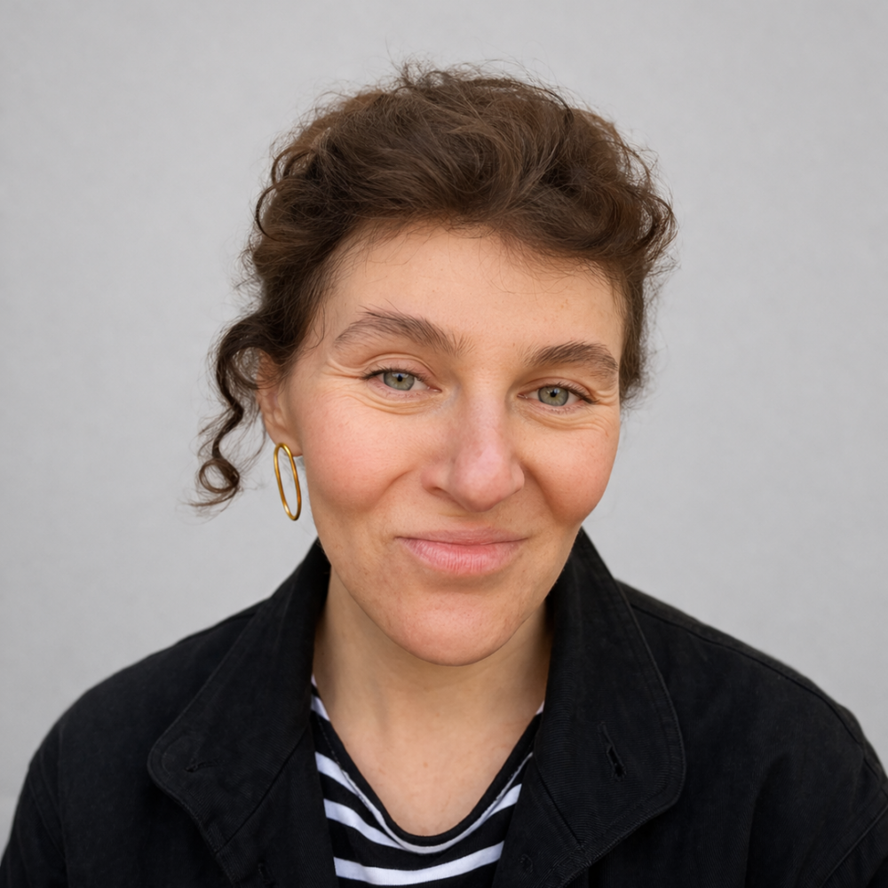 A woman with curly brown hair and blue eyes is smiling softly. She is wearing a black jacket over a black and white striped shirt and gold hoop earrings, with a plain light gray background.