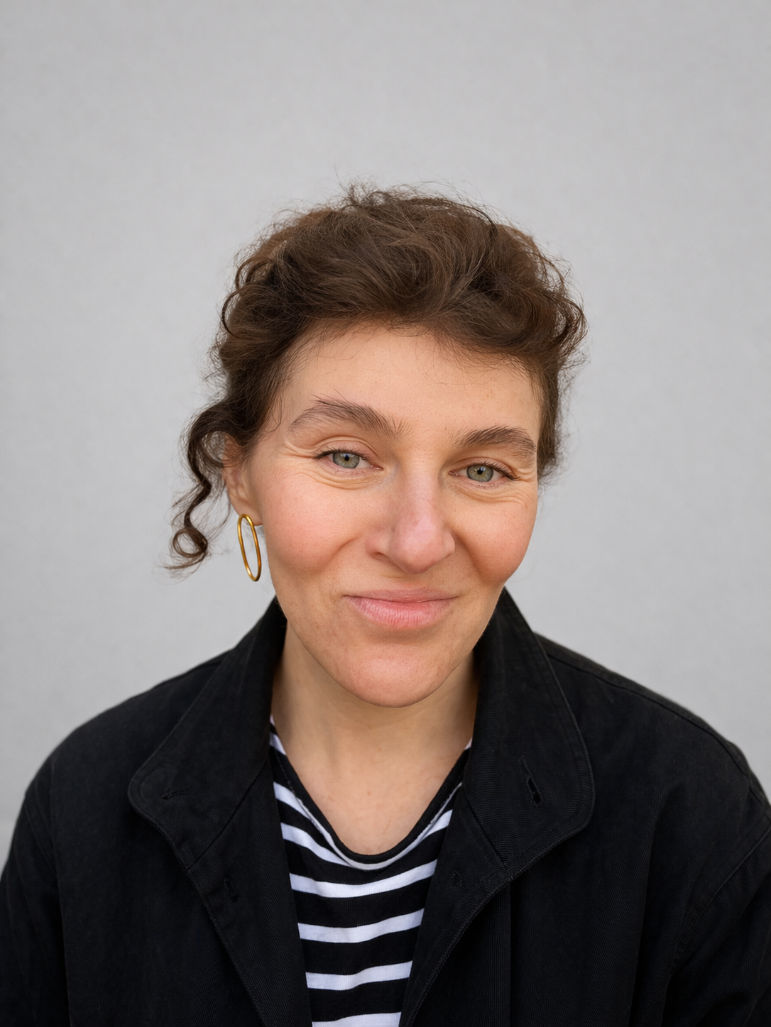 A woman with curly brown hair and blue eyes is smiling softly. She is wearing a black jacket over a black and white striped shirt and gold hoop earrings, with a plain light gray background.