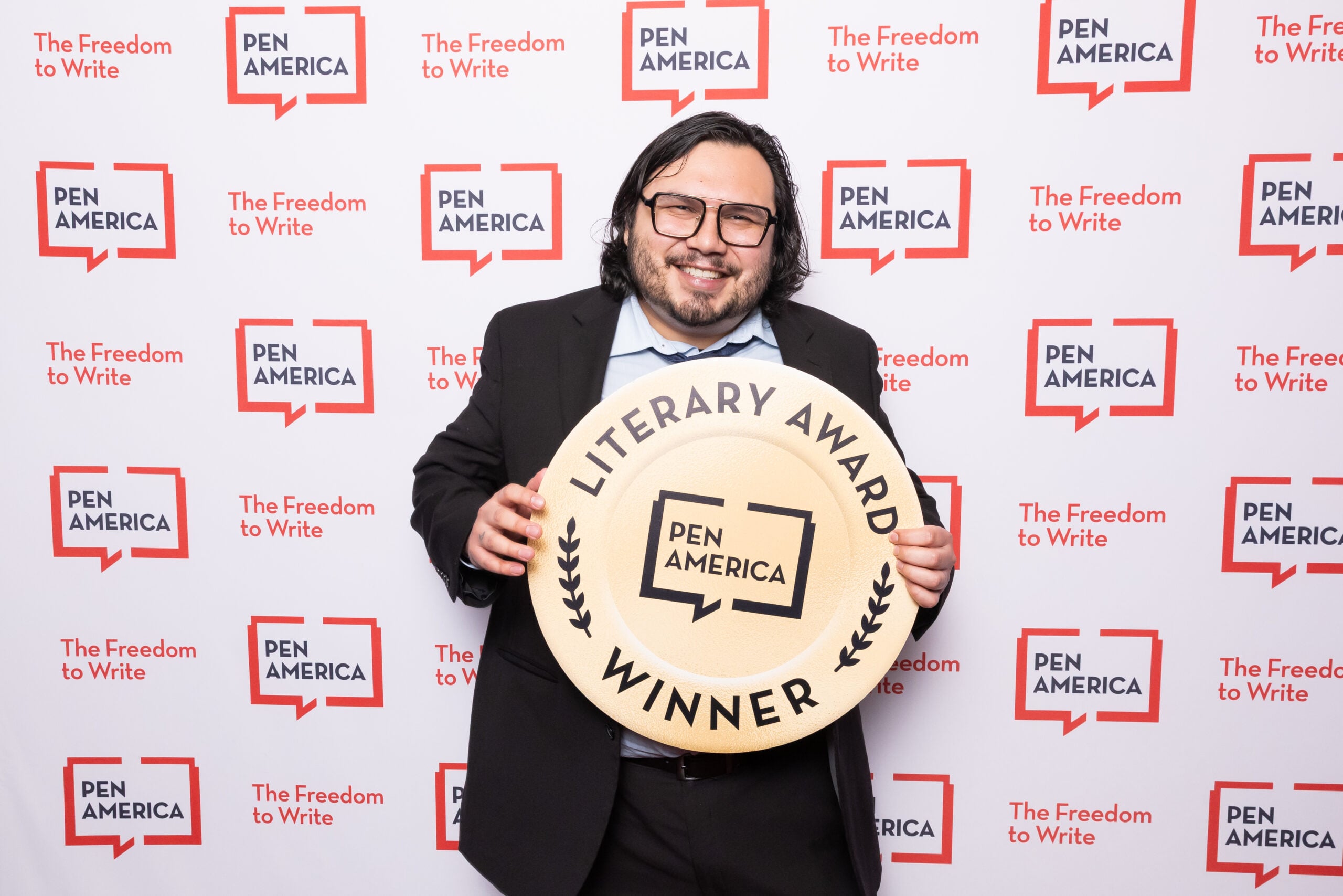 A smiling man in a suit stands in front of a PEN America backdrop, holding a large circular sign that reads PEN America Literary Award Winner.