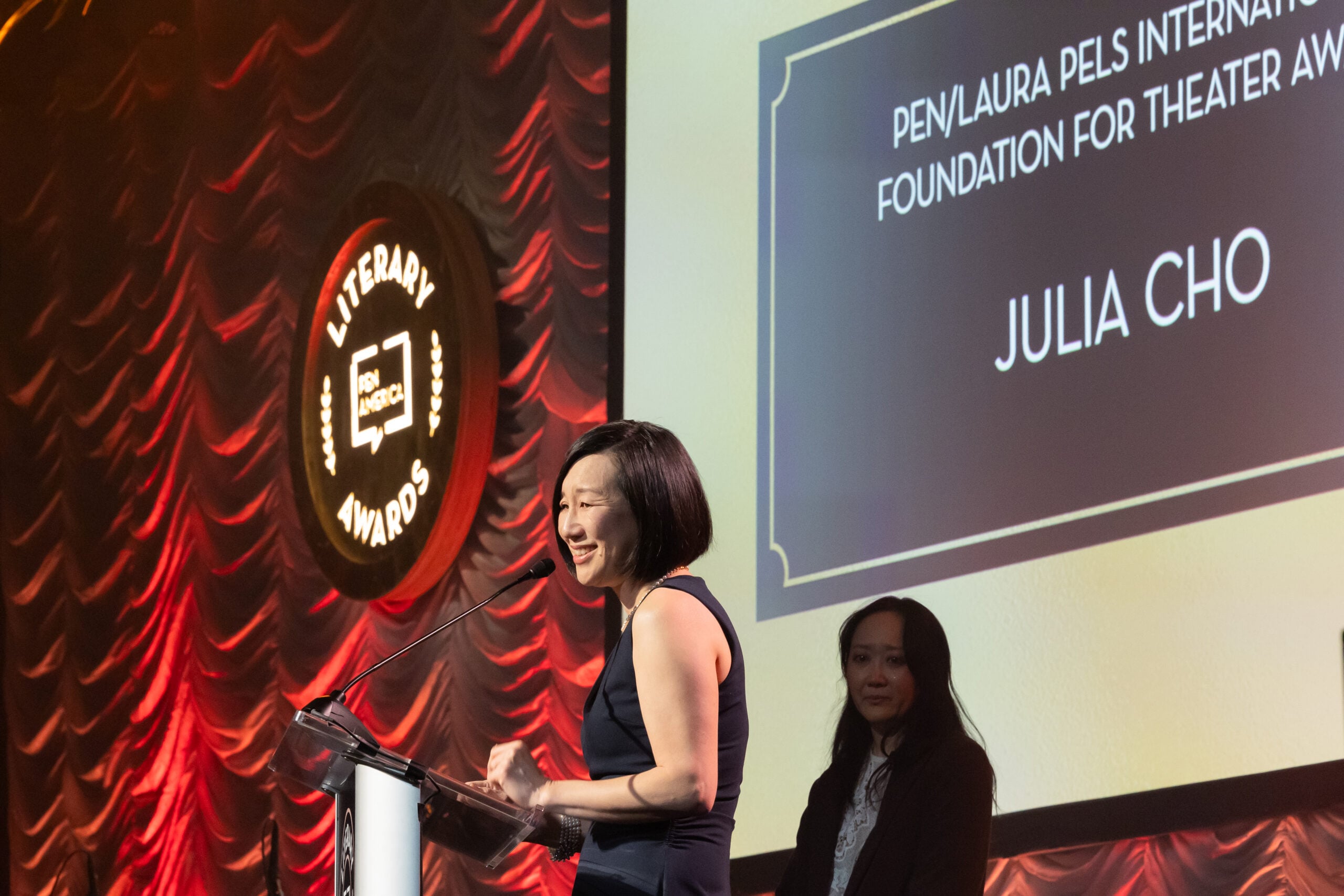 A woman stands at a podium smiling while speaking on stage at a literary awards event, with another woman standing behind her. The screen shows PEN/Laura Pels International Foundation for Theater Award: Julia Cho.