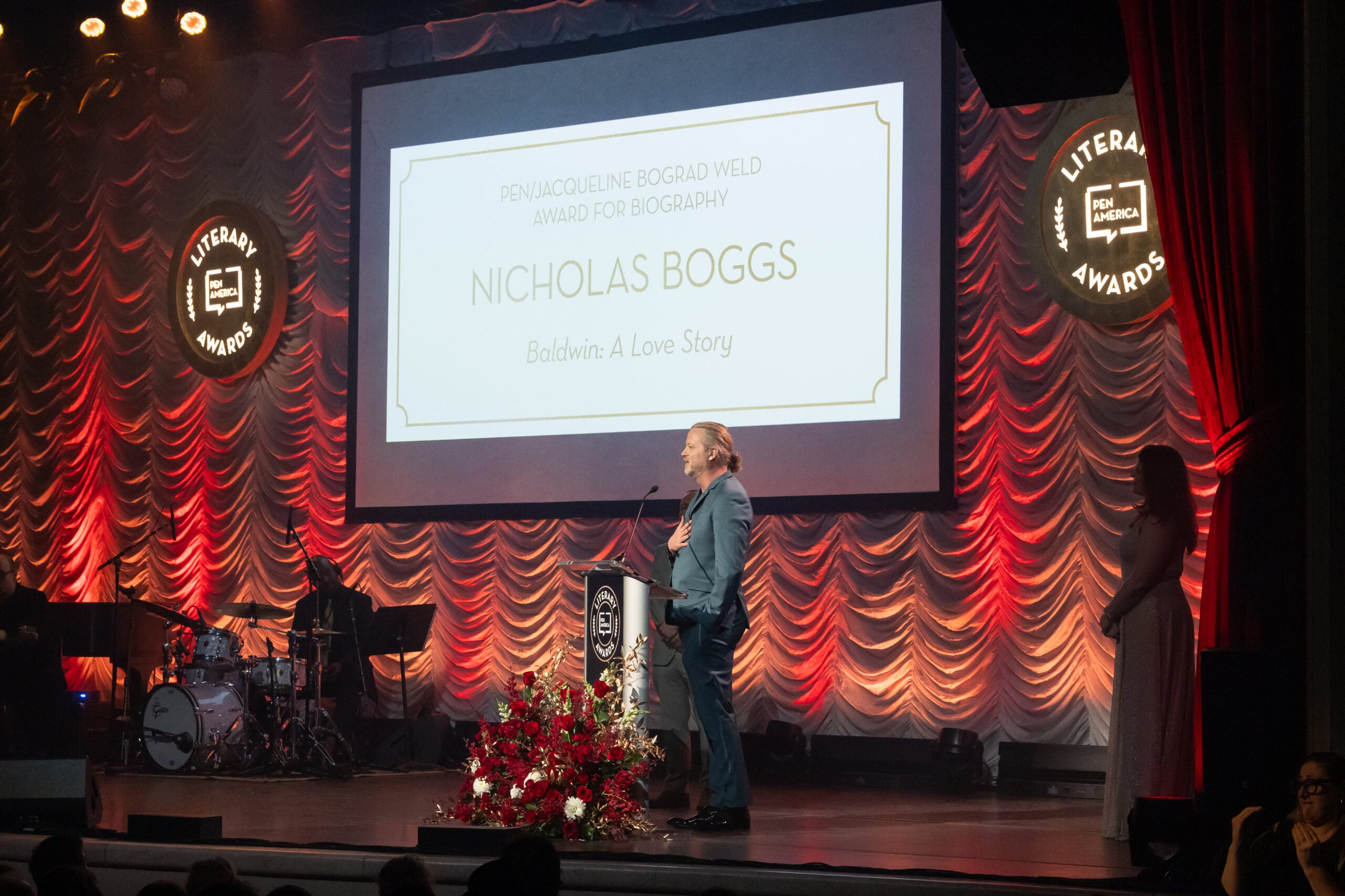 A person stands at a podium on stage at an awards ceremony. Behind them, a screen displays Nicholas Boggs, Baldwin: A Love Story as the winner of the PEN/Jacqueline Bograd Weld Award for Biography. A band and another person are also on stage.