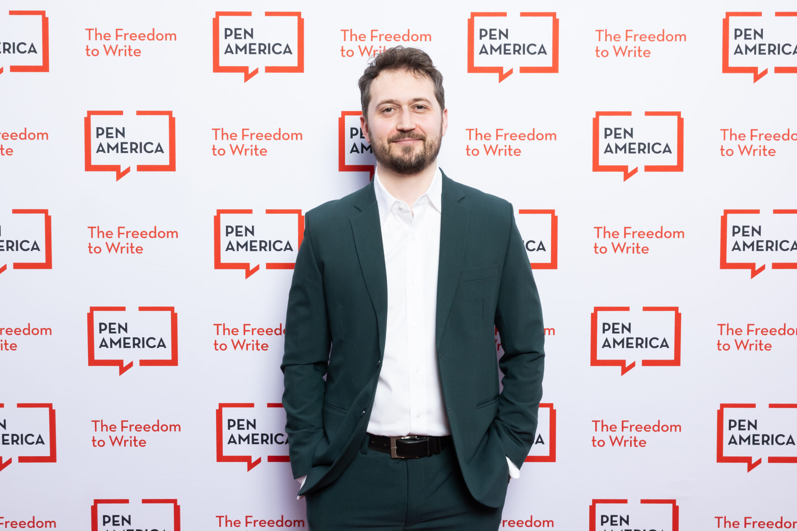 A man in a dark green suit with a white shirt stands smiling with hands in his pockets in front of a step-and-repeat backdrop with PEN America and The Freedom to Write logos.