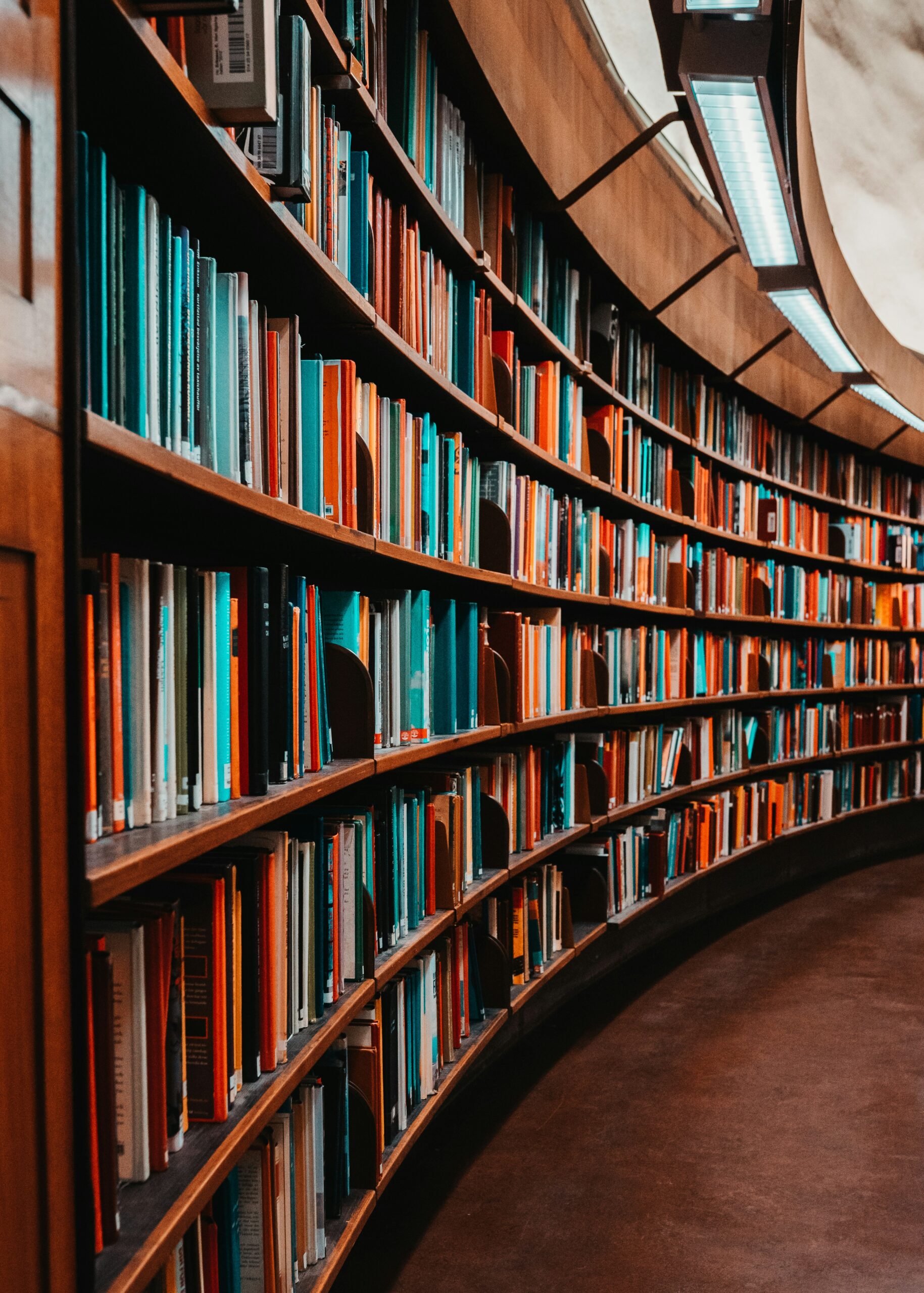 Curved wooden bookshelves filled with colorful books line a corridor in a library, illuminated by overhead lights. The scene creates a warm and inviting atmosphere for reading and study.