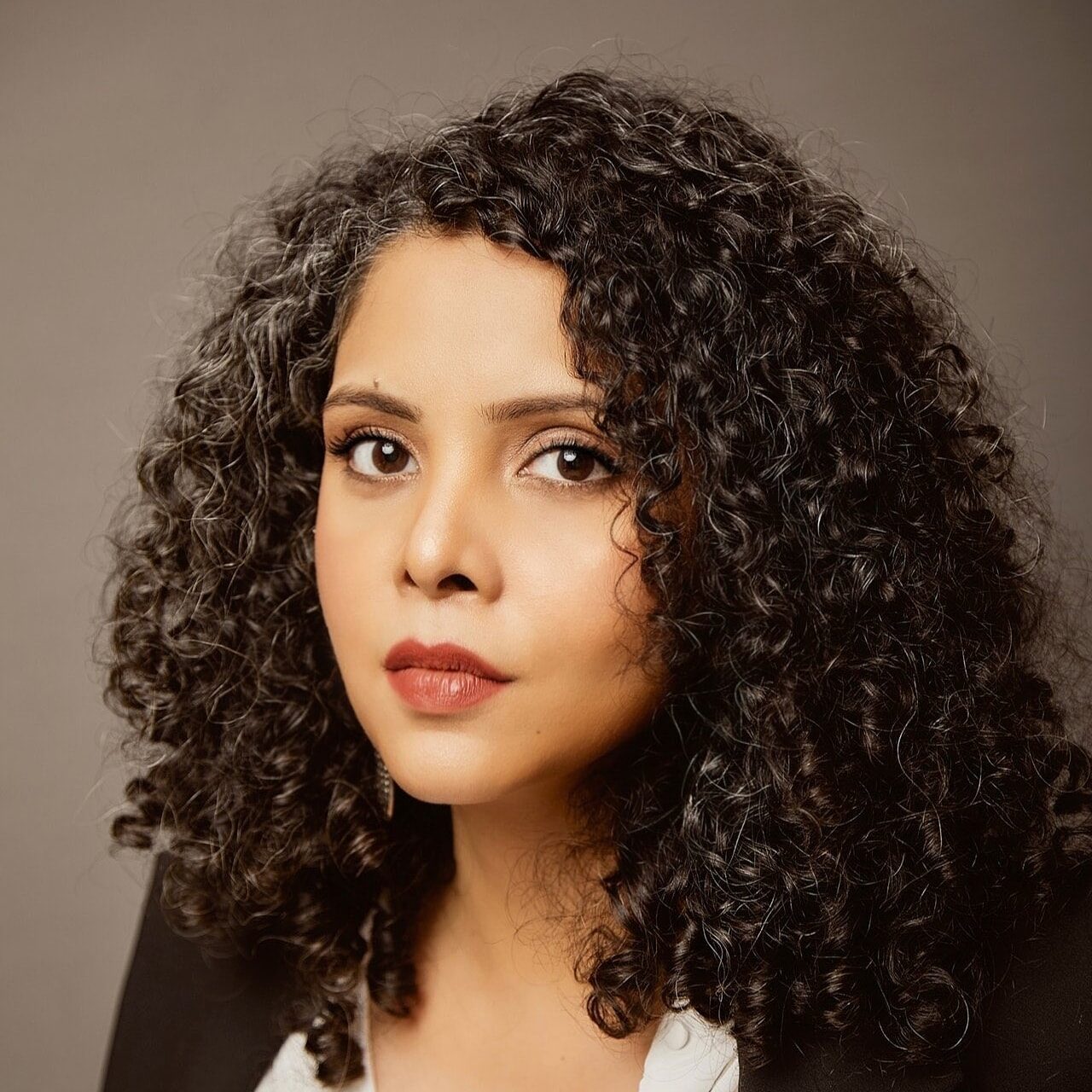 A woman with curly dark hair and a few gray strands, wearing a white blouse and a black blazer, looks directly at the camera with a neutral expression against a plain background.