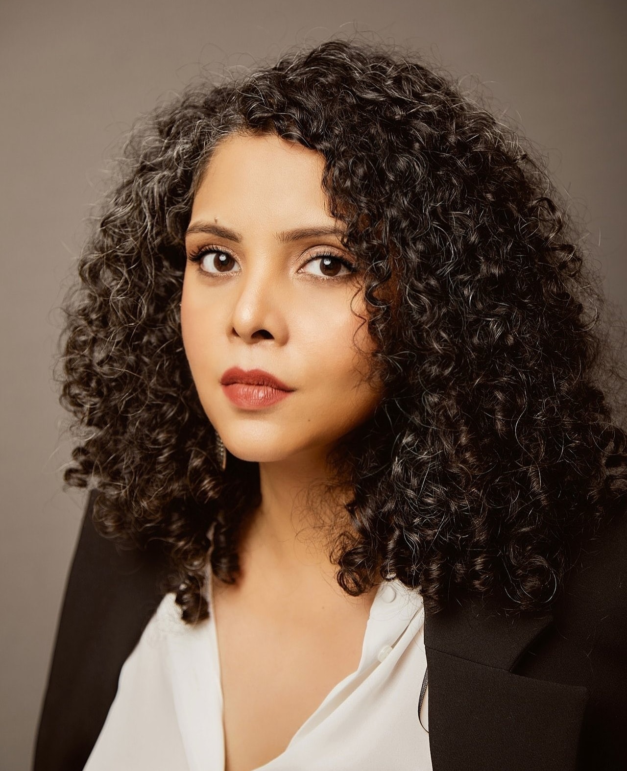 A woman with curly dark hair and a few gray strands, wearing a white blouse and a black blazer, looks directly at the camera with a neutral expression against a plain background.