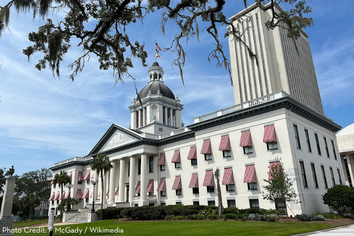 The image shows the Florida State Capitol building in Tallahassee, featuring a white facade, red-and-white striped window awnings, a central dome, and a tall modern tower in the background under a blue sky.