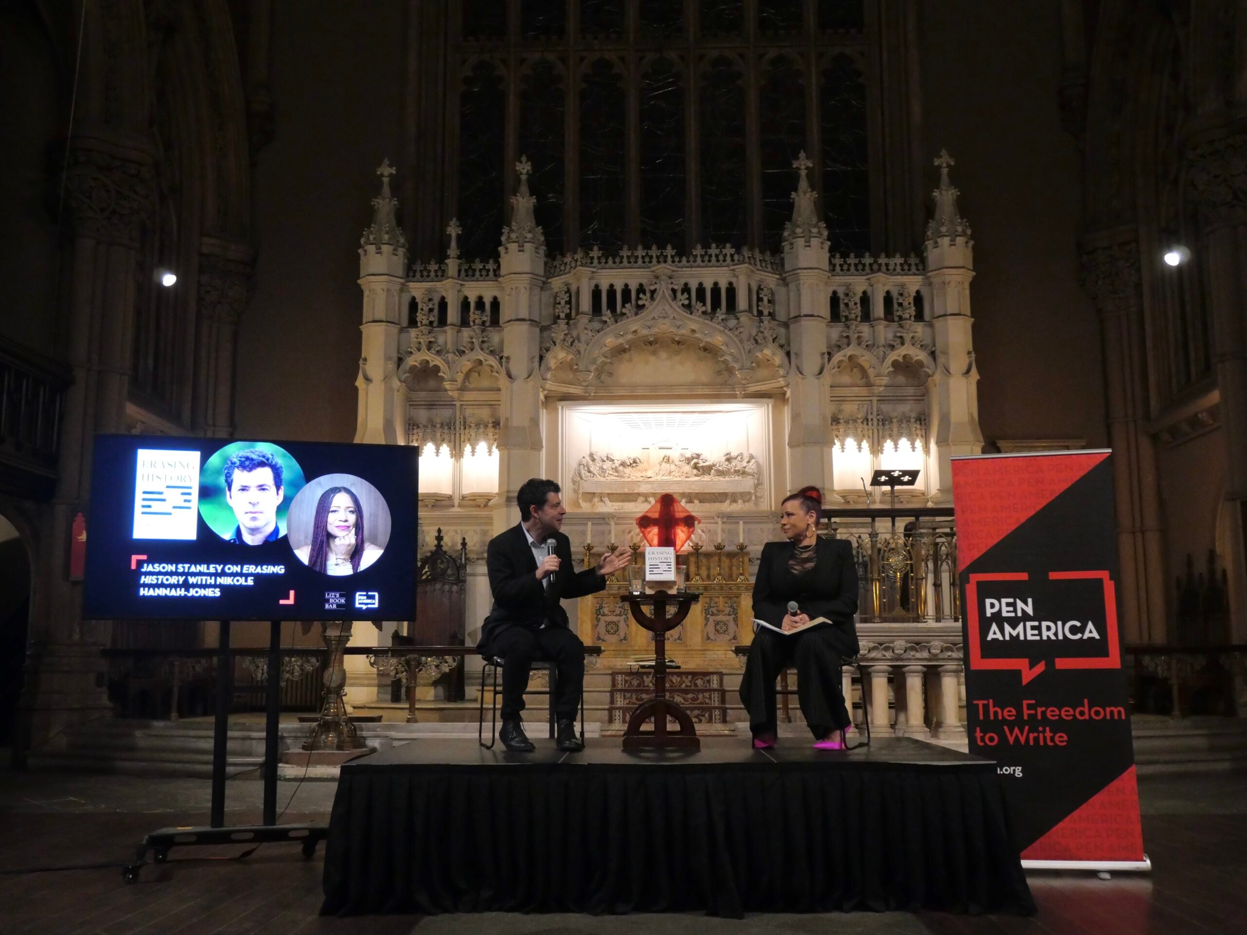 Two people sit and speak on a stage in front of an ornate church altar. A screen shows photos and names of two individuals. A PEN America banner reads “The Freedom to Write.” An audience is implied off-camera.