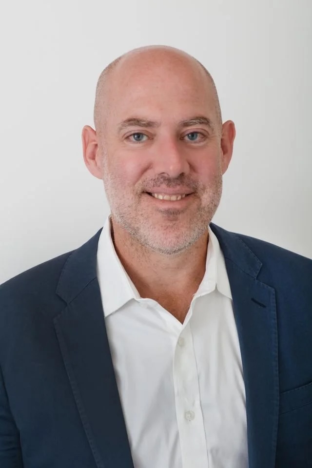 A man with a shaved head and light stubble is smiling at the camera. He is wearing a white collared shirt and a dark blue blazer, standing in front of a plain white background.