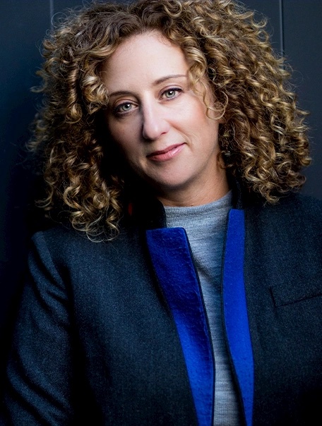 A woman with curly light brown hair, wearing a dark jacket with a blue collar and a gray top, stands in front of a dark background, smiling softly at the camera.