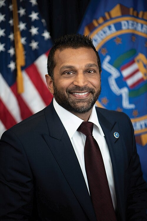 A man in a dark suit, white shirt, and red tie smiles at the camera. He stands in front of U.S. and FBI flags, with a lapel pin visible on his jacket.