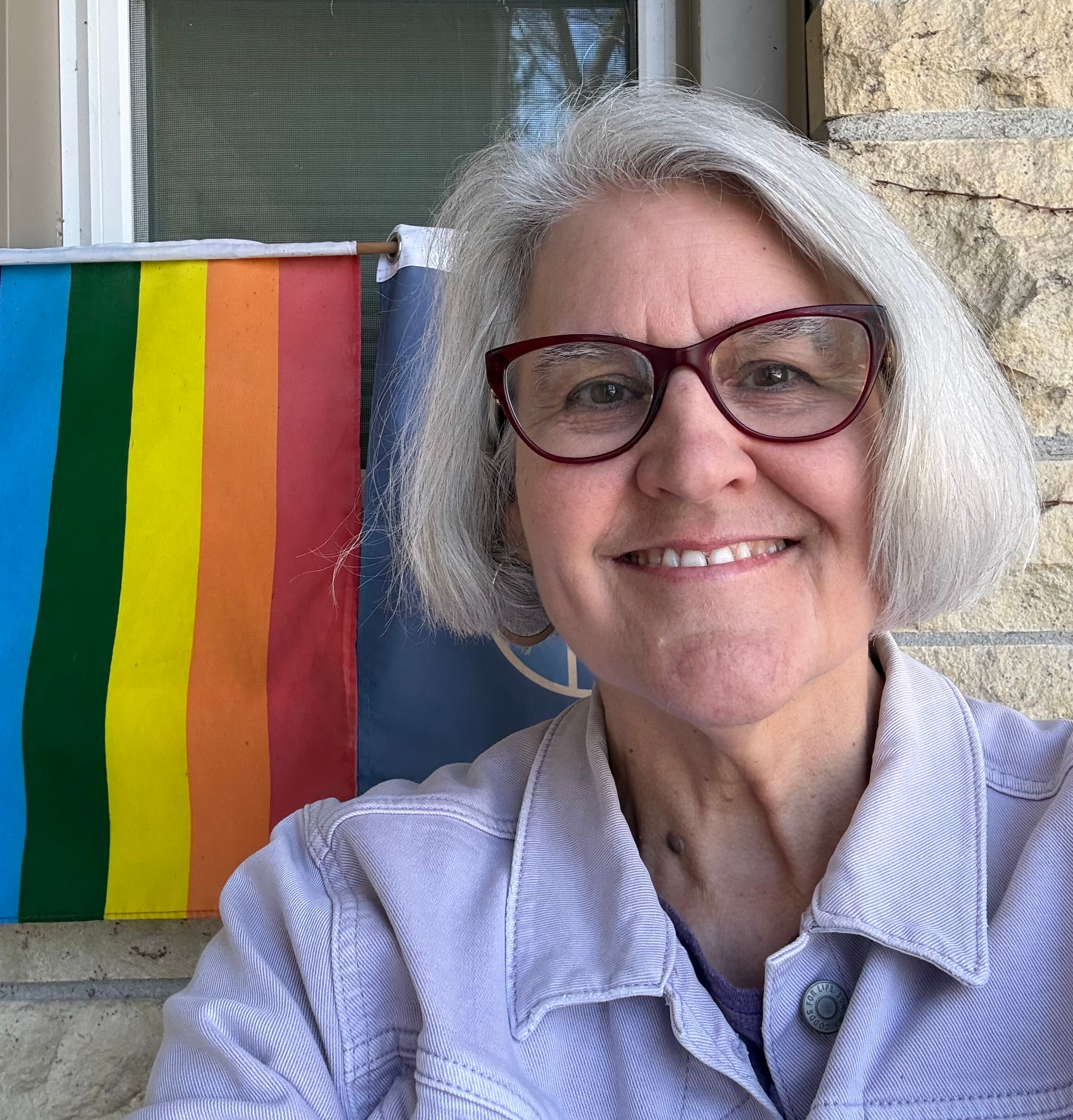 A smiling person with short gray hair and red glasses poses outside near a rainbow pride flag hanging on a wall. Stone and part of a window are visible in the background.