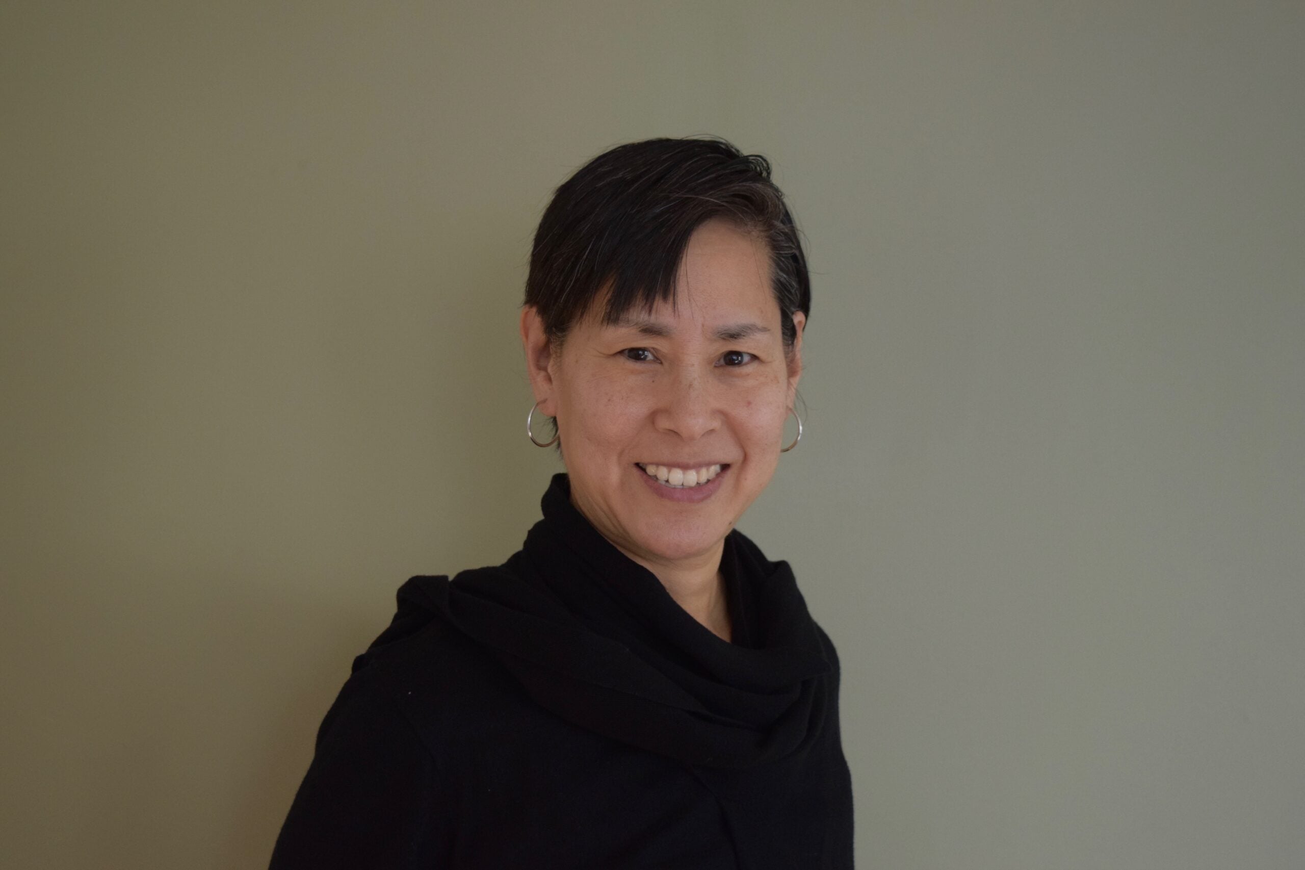 A person with short dark hair, wearing hoop earrings and a black top, smiling and standing in front of a plain light-colored wall.