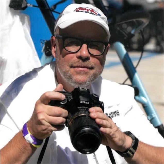 A person wearing glasses, a white cap, and a white shirt is holding a Nikon camera and looking at the lens, with a blue bicycle and blurred background behind them.