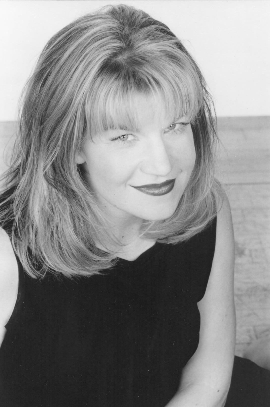 A woman with shoulder-length, light-colored hair and bangs smiles slightly while looking up at the camera. She wears a sleeveless dark top and has light makeup. The photo is in black and white.
