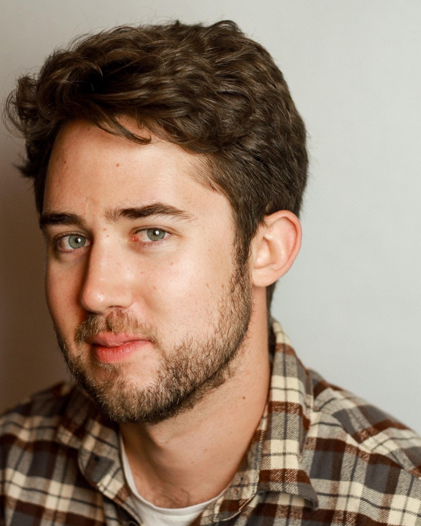 A young man with short brown hair and a beard, wearing a plaid shirt, looks slightly to the side with a neutral expression against a plain background.