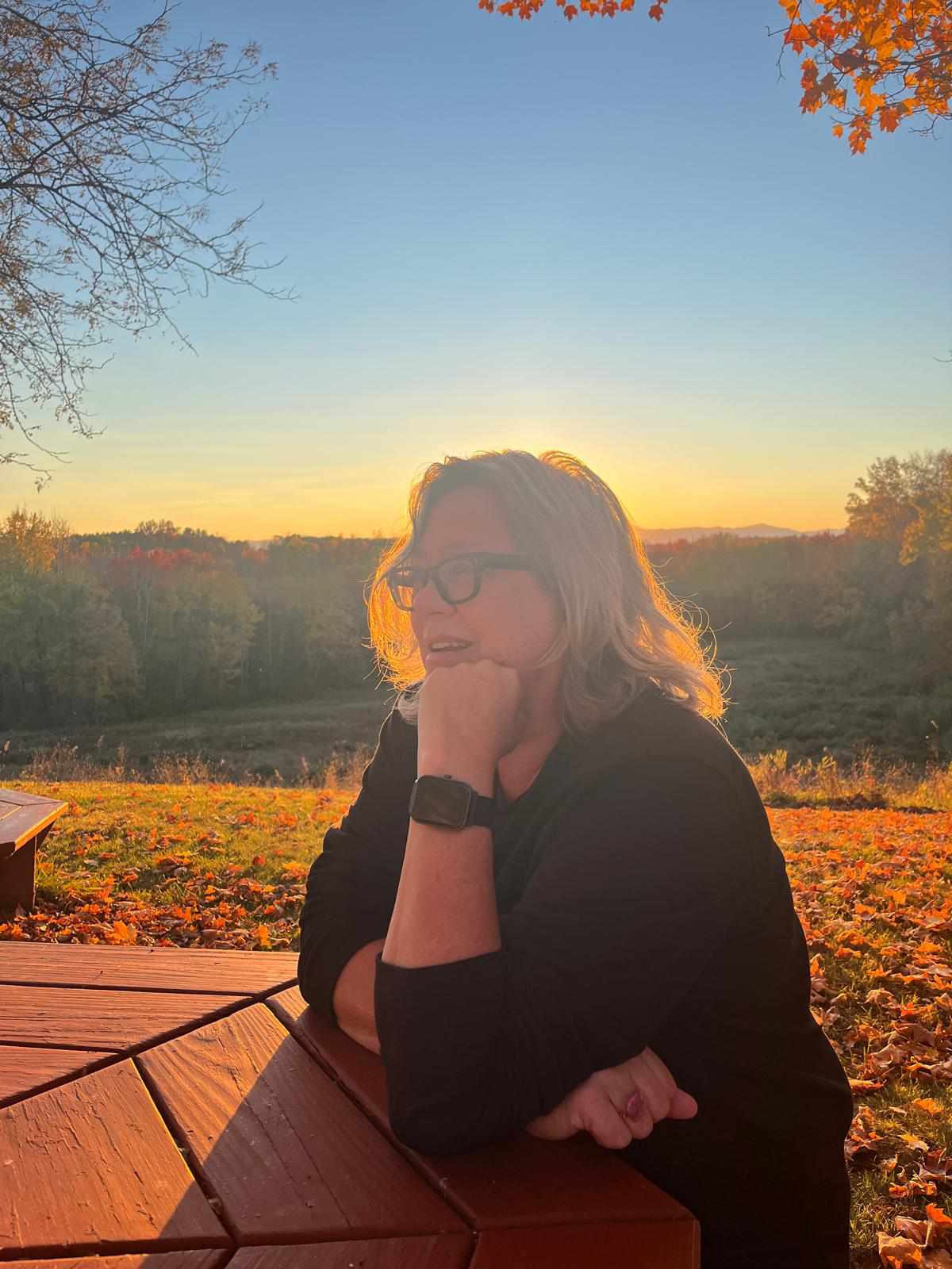A person with light hair and glasses sits at a wooden picnic table, resting their chin on their hand, with a scenic autumn landscape and a setting sun in the background.