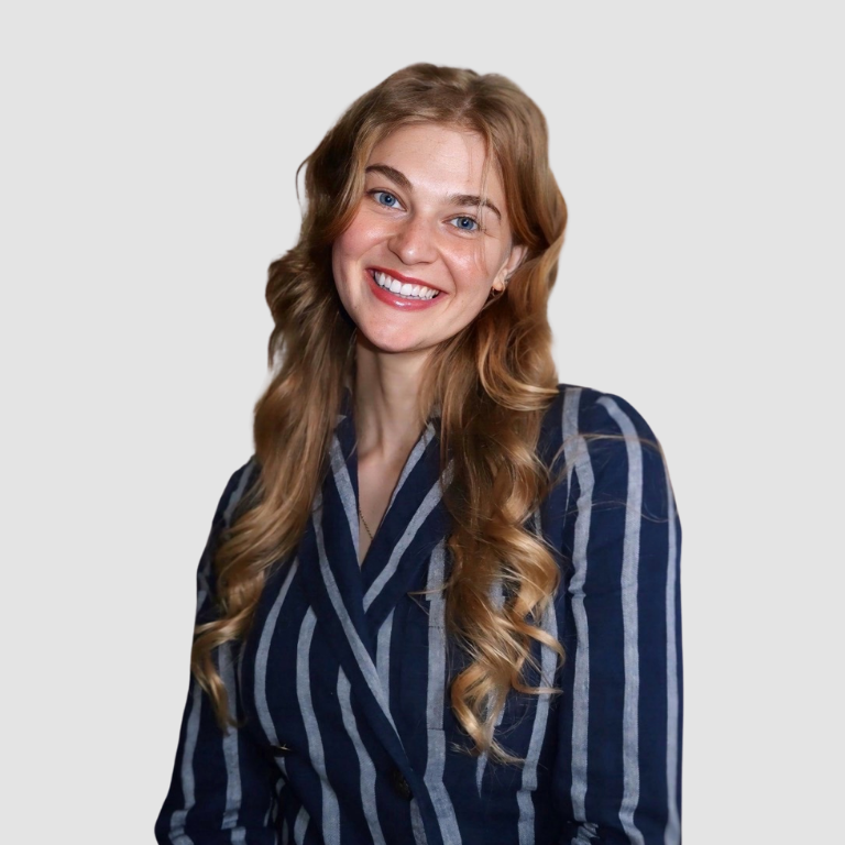 A smiling woman with long, wavy blonde hair wearing a navy blue blazer with white vertical stripes, standing against a plain light gray background.