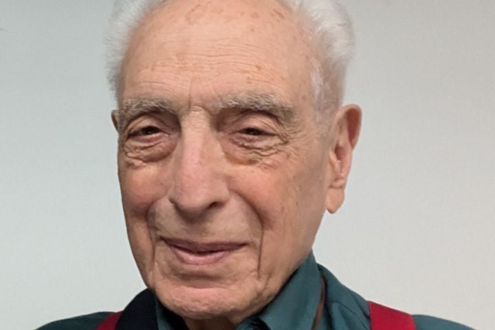 An elderly man with short white hair and fair skin is smiling gently. He is wearing a teal shirt and red suspenders, standing against a plain, light-colored background.