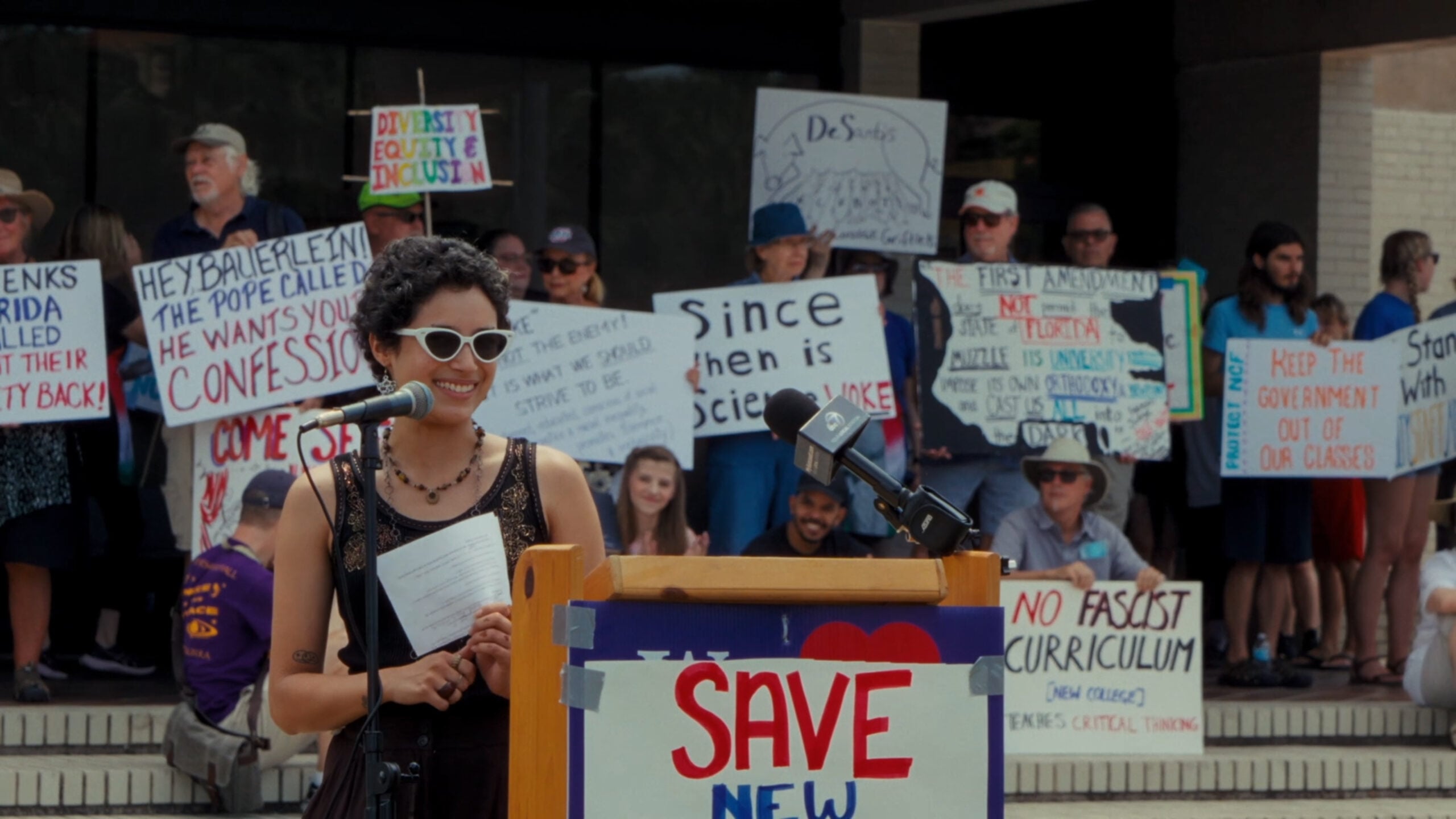 A woman with curly hair and white sunglasses speaks at a podium with a “SAVE NEW” sign, while people behind her hold protest signs about science, education, and curriculum.