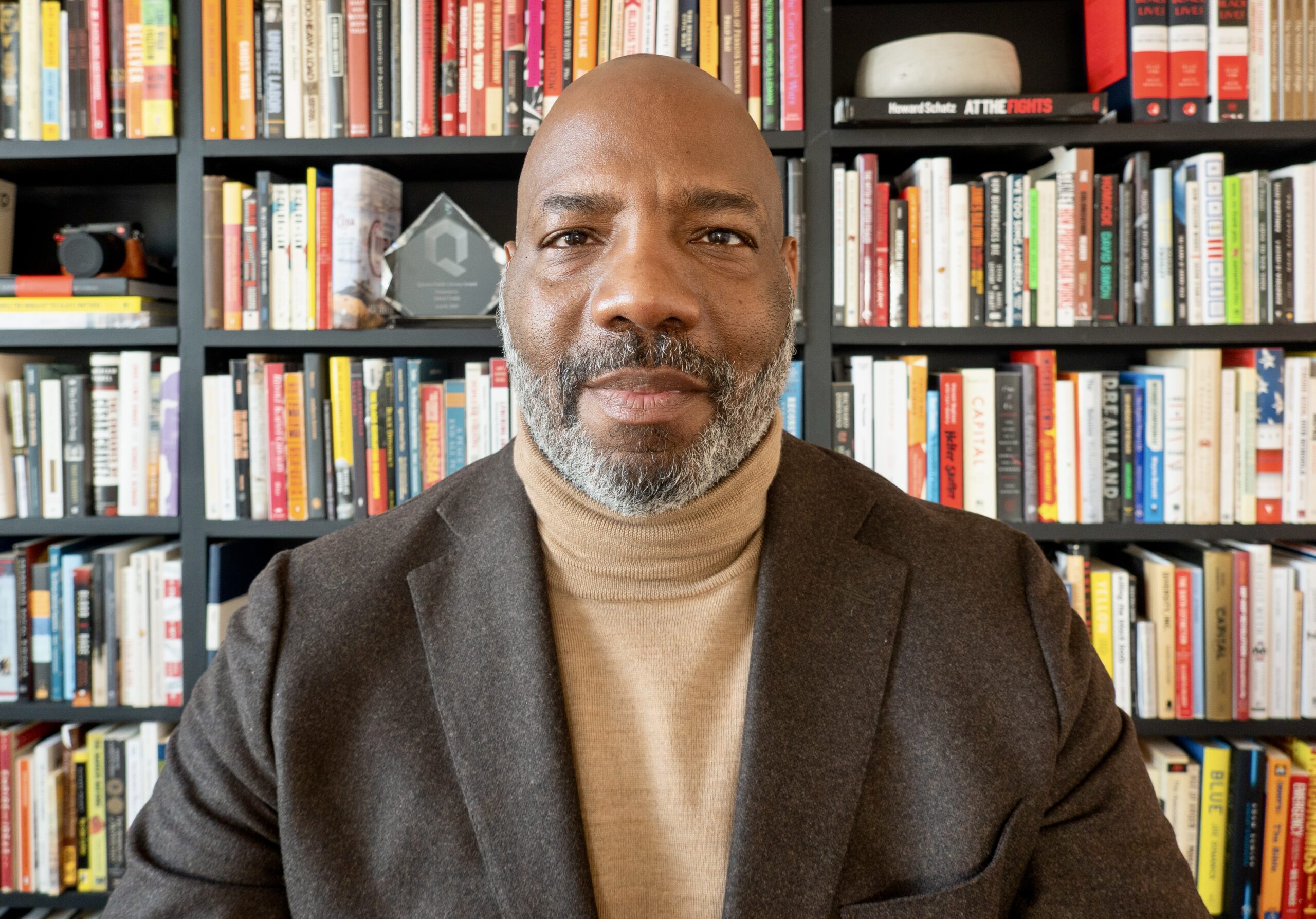 A man with a shaved head and gray beard, wearing a brown blazer and tan turtleneck, sits in front of a bookshelf filled with colorful books.
