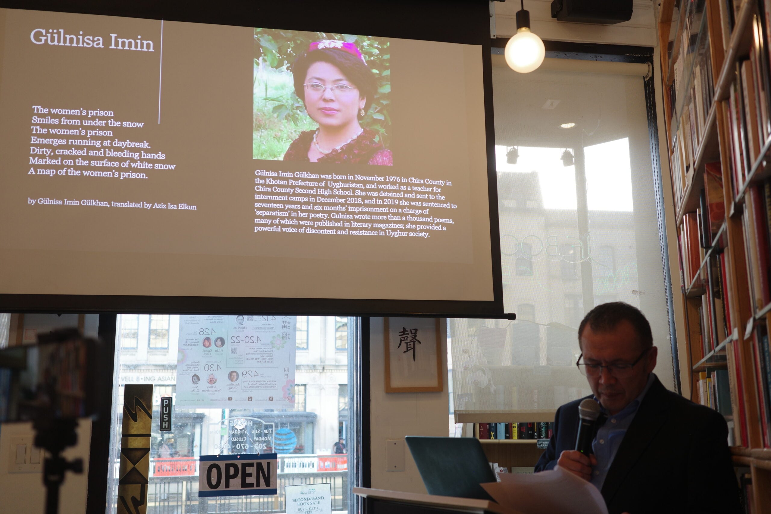 A man reads at a podium in a bookstore, while a projected screen displays a photo and biography of poet Gülnisa Imin, along with her poem “The women’s prison” and a translation credit. Shelves of books and a window are visible.