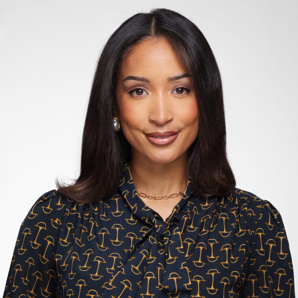 A woman with straight dark hair and medium skin tone smiles slightly. She wears a navy blouse with a gold pattern, gold necklace, and pearl earrings, posing against a plain white background.
