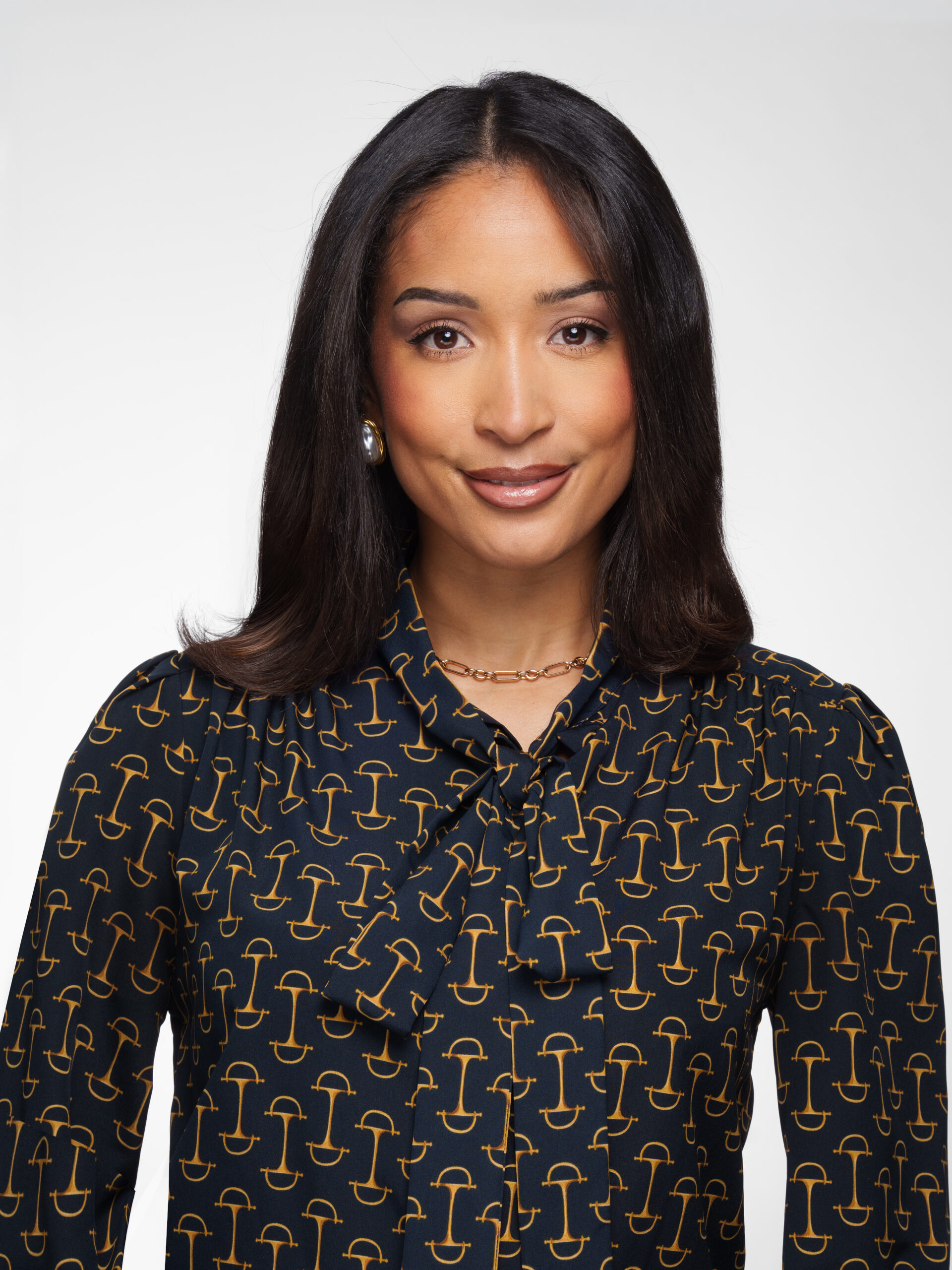 A woman with straight dark hair and medium skin tone smiles slightly. She wears a navy blouse with a gold pattern, gold necklace, and pearl earrings, posing against a plain white background.