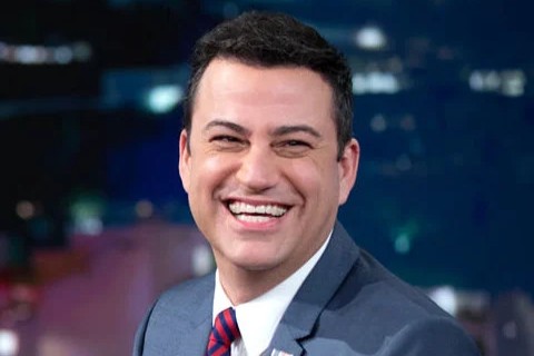 A man in a suit and tie is smiling broadly while sitting indoors, with a blurred, colorful background.
