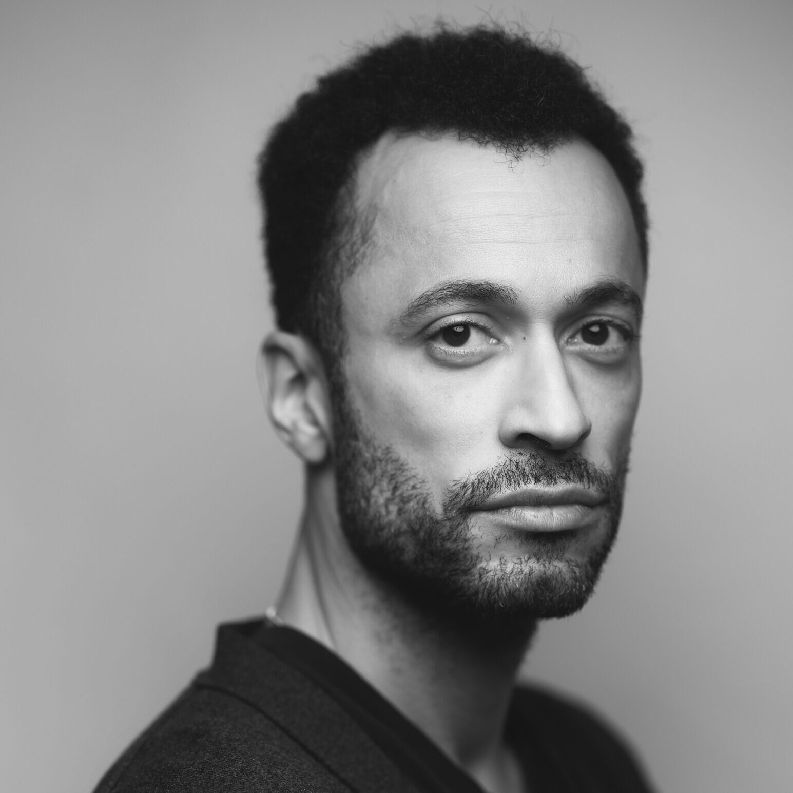 Black and white portrait of a man with short curly hair and a beard, wearing a dark jacket over a shirt, looking seriously at the camera against a plain background.