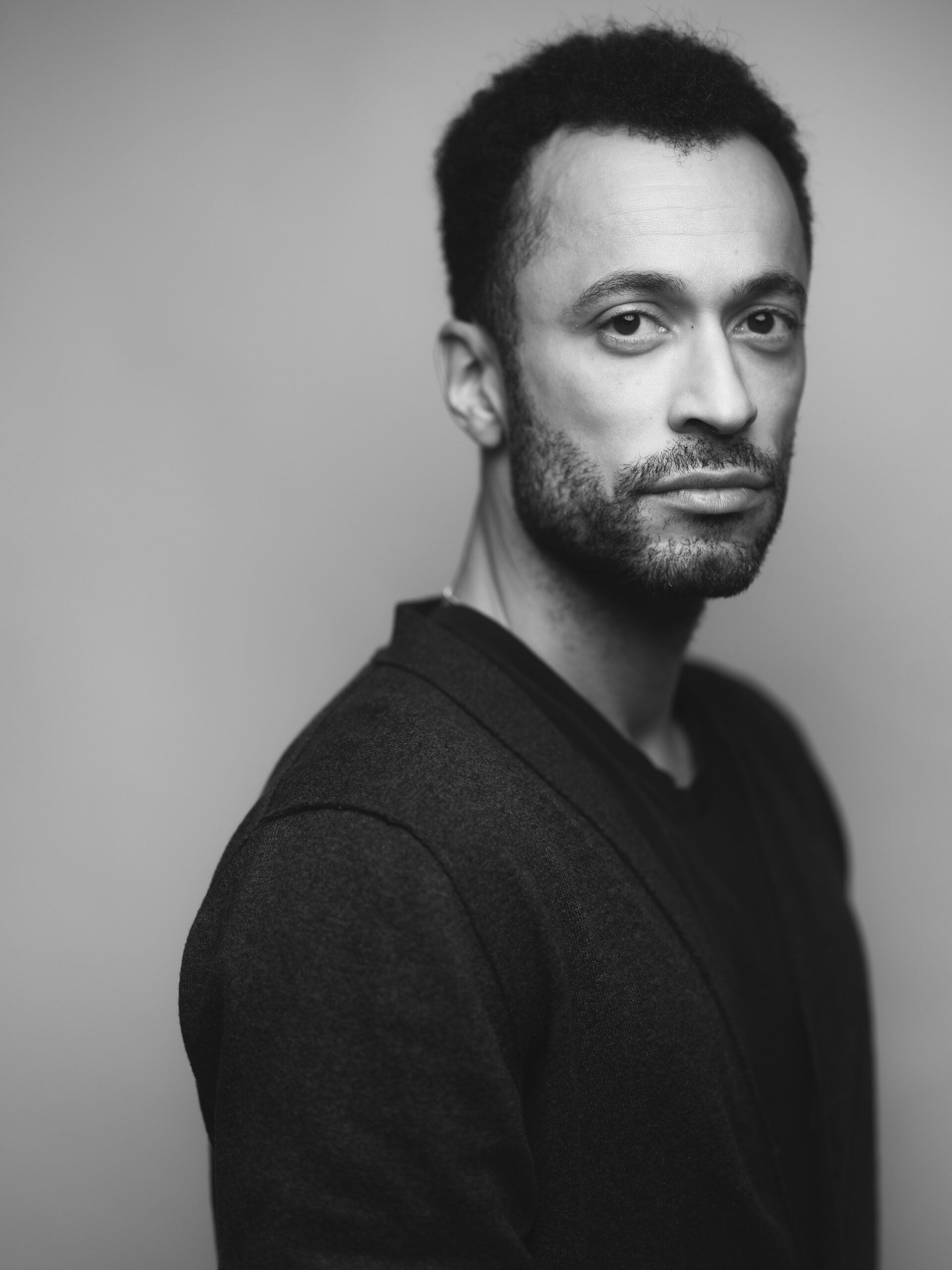 Black and white portrait of a man with short curly hair and a beard, wearing a dark jacket over a shirt, looking seriously at the camera against a plain background.