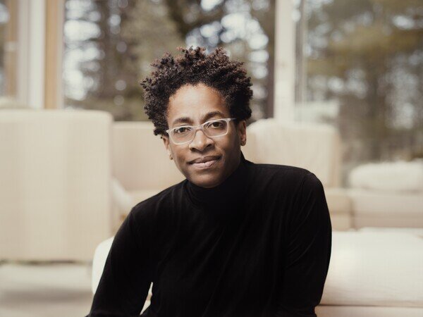 A person with short curly hair and glasses, wearing a black turtleneck, sits indoors in front of large windows with a blurred background of trees and neutral-toned furniture.