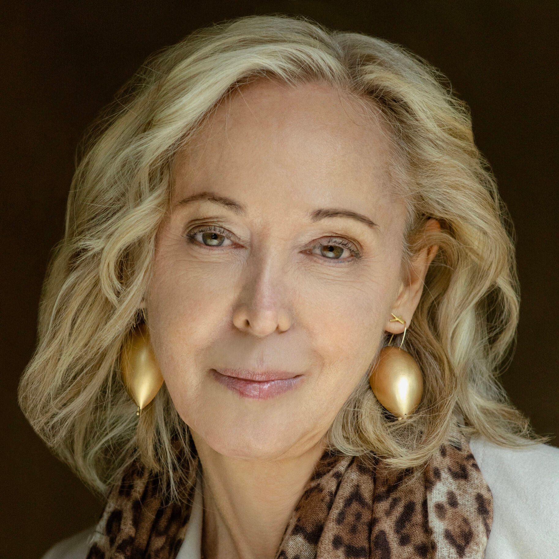 A woman with wavy blonde hair, wearing large gold earrings, a light-colored jacket, and a leopard-print scarf, smiles softly against a dark background.