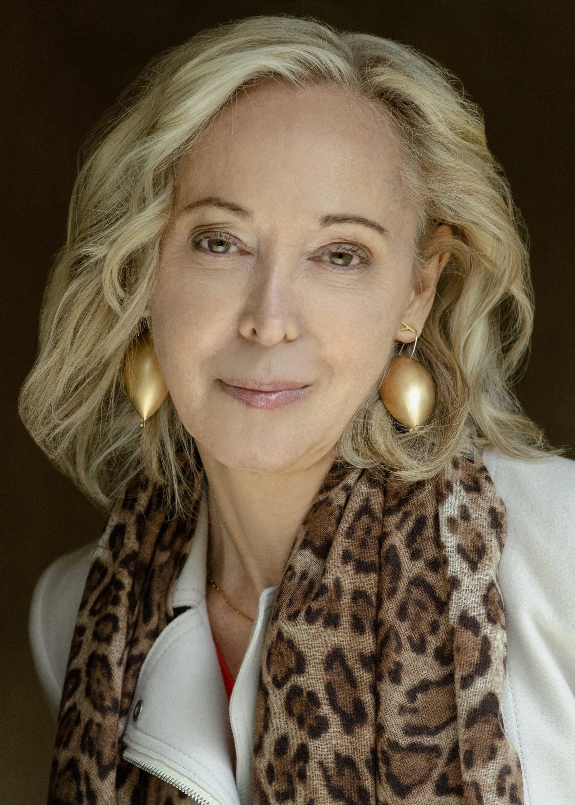 A woman with wavy blonde hair, wearing large gold earrings, a light-colored jacket, and a leopard-print scarf, smiles softly against a dark background.