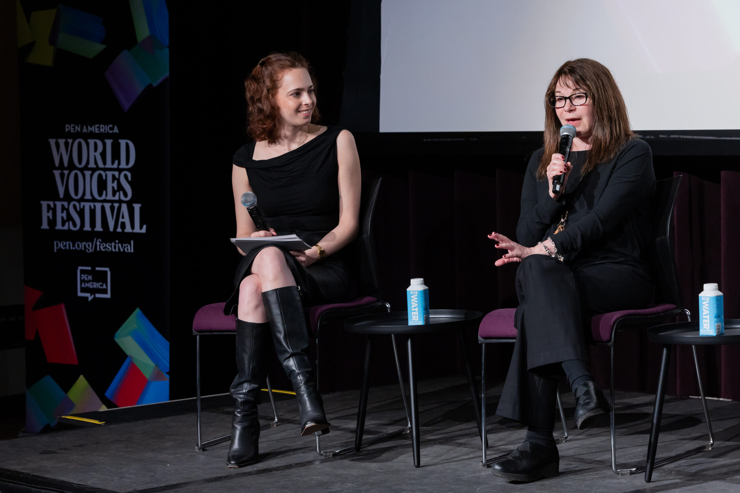 Two women sit on stage at the PEN America World Voices Festival. One holds a microphone and is speaking while the other listens attentively, holding a notecard and microphone. Both have water cartons on the table beside them.