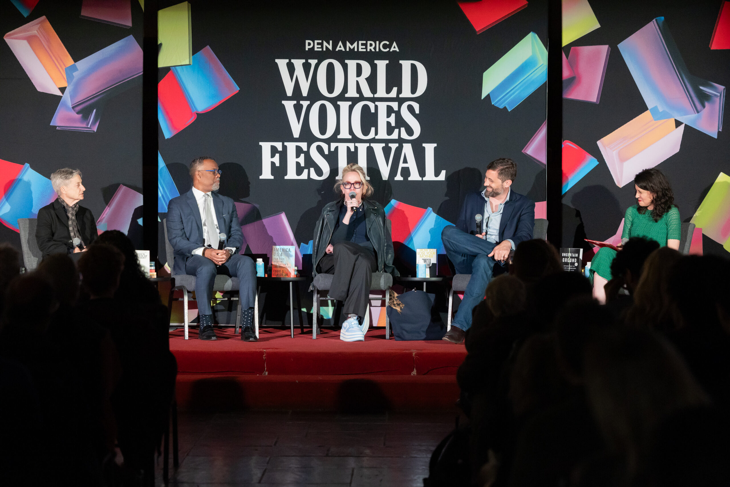 Five panelists sit onstage speaking to an audience at the PEN America World Voices Festival, with a colorful backdrop featuring the event’s name and abstract book designs.