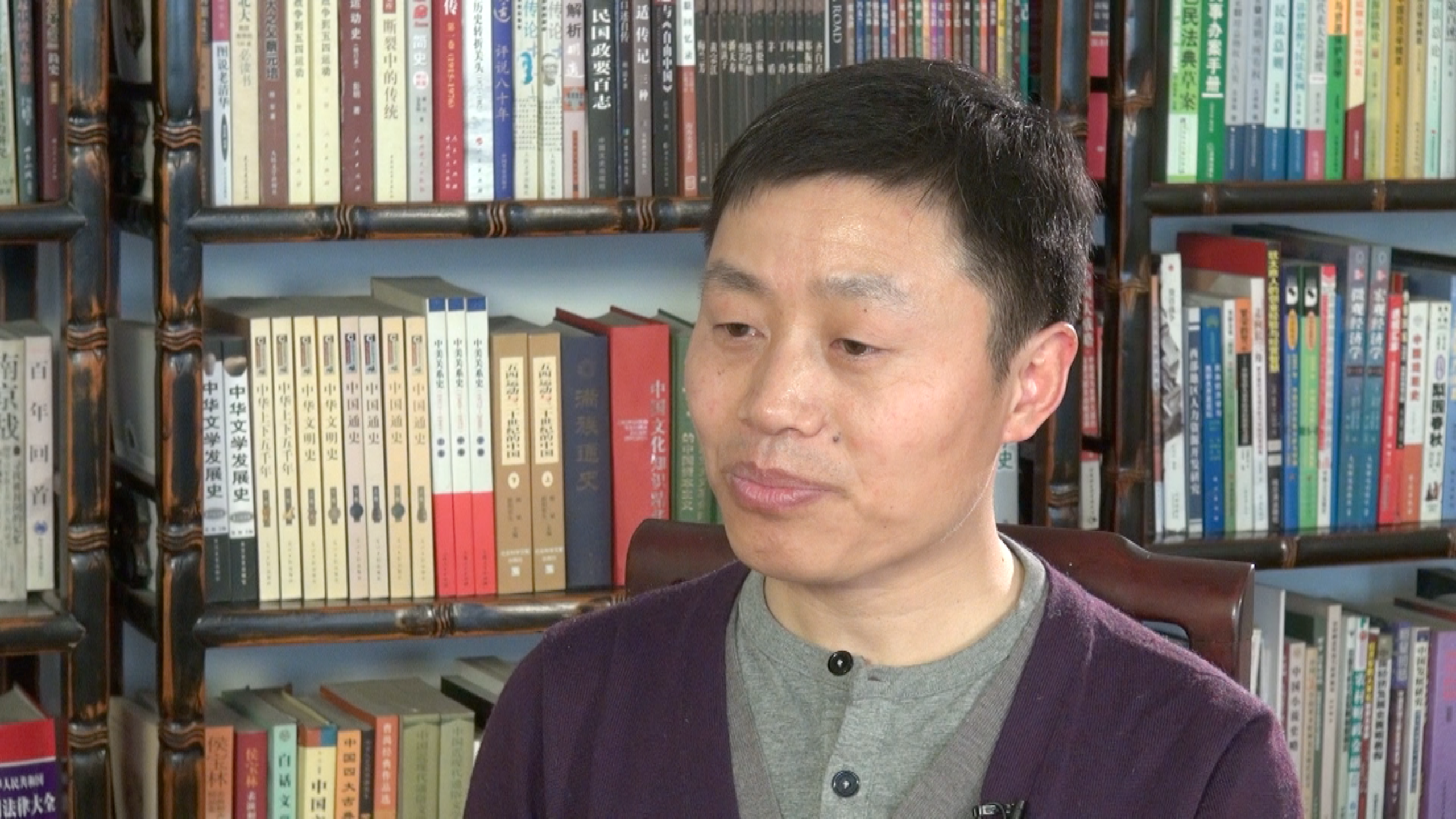 A man in a purple cardigan and grey shirt sits in front of bookshelves filled with books, looking slightly to the side with a neutral expression.