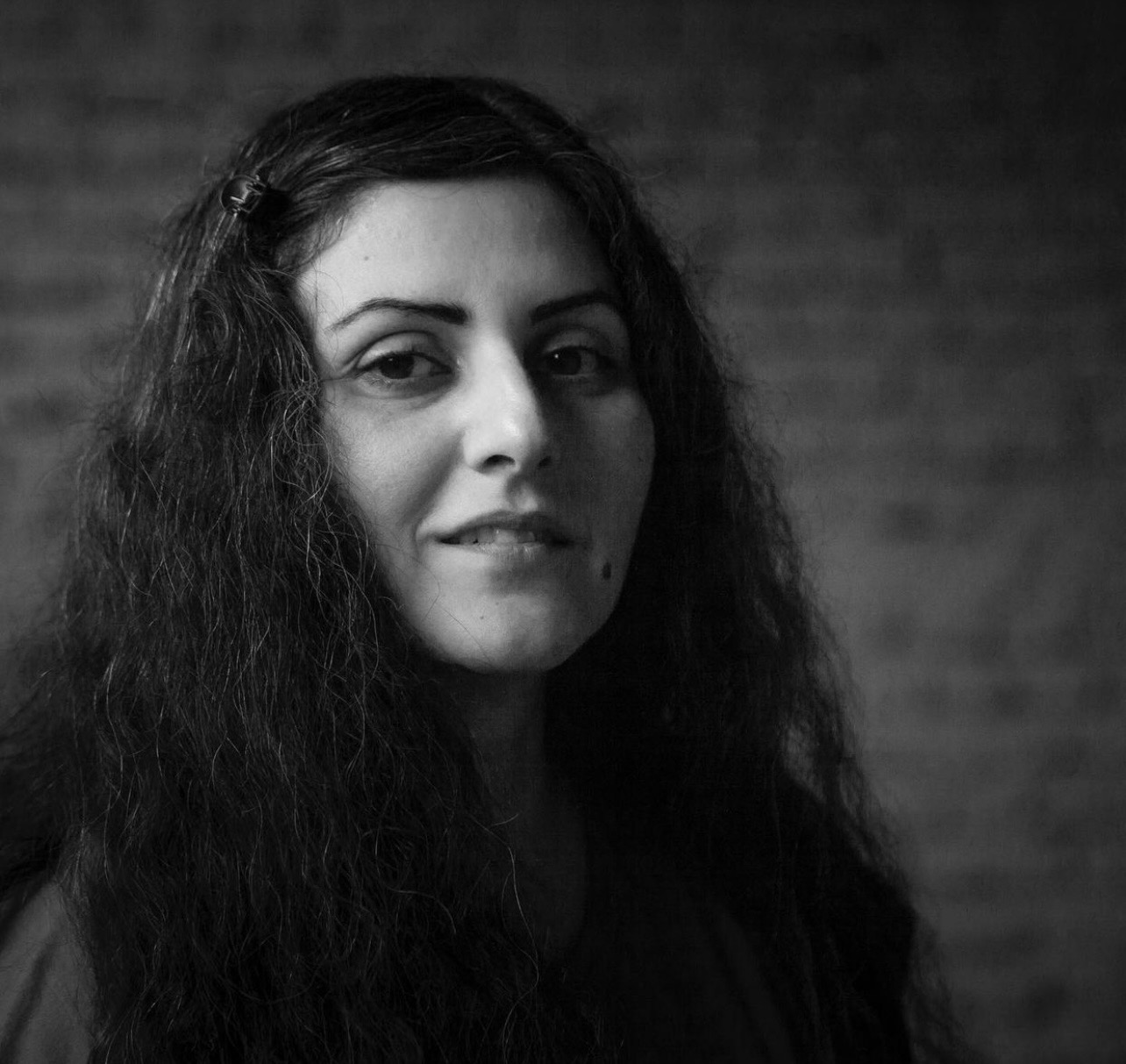 Black and white portrait of a woman with long wavy hair, a small mole on her cheek, and a slight smile, standing in front of a blurred brick wall background.