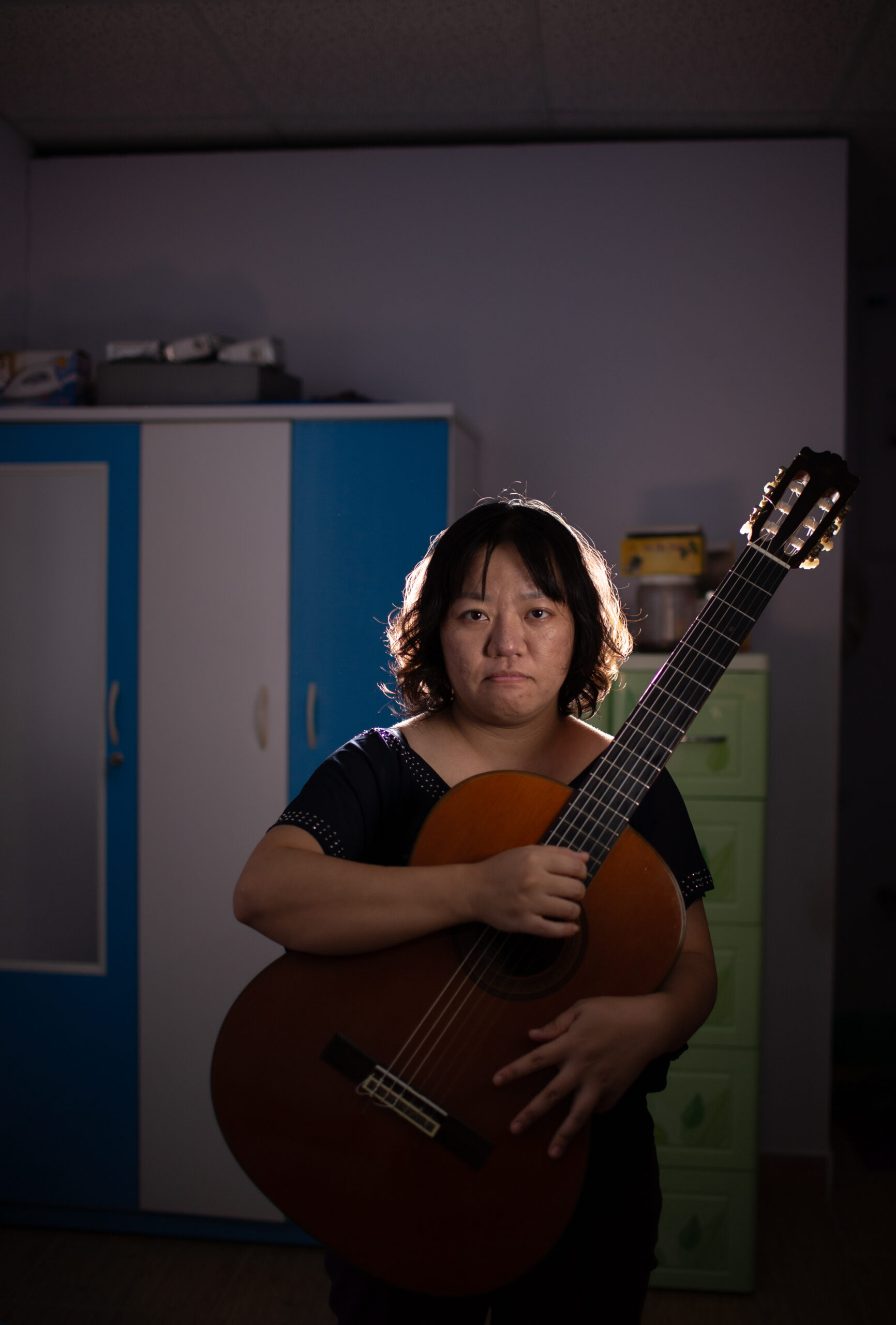A woman with short hair stands indoors holding a classical guitar. She is facing the camera with a neutral expression. There is a blue and white cabinet and a green drawer unit in the background.