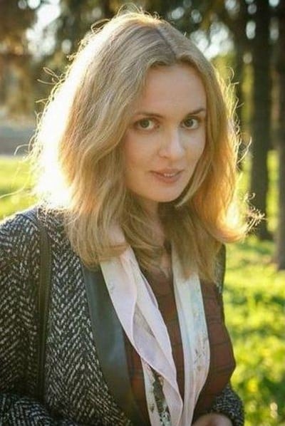 A woman with shoulder-length blonde hair stands outdoors in sunlight, wearing a patterned jacket and a light scarf, with trees and greenery blurred in the background.