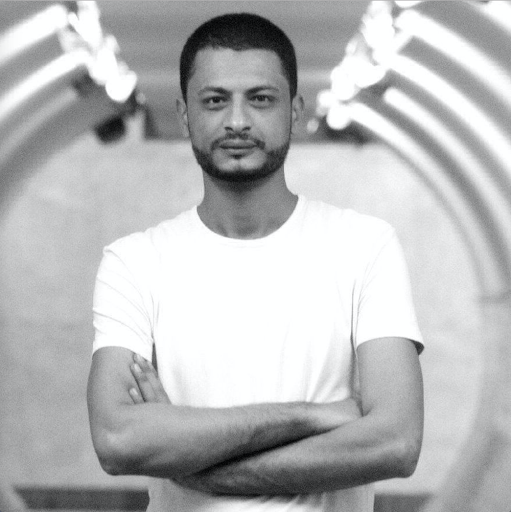 A man in a white t-shirt stands with arms crossed in a tunnel, looking at the camera. The photo is black and white, and the tunnel features curved lights along the ceiling.