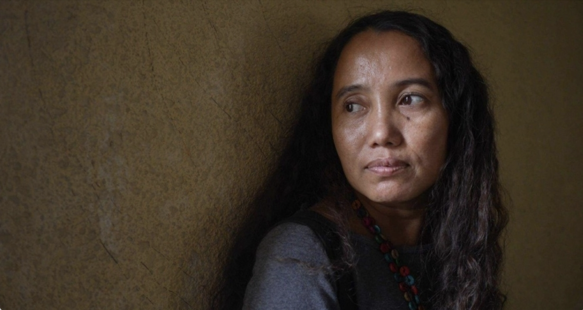 A woman with long dark hair and a necklace looks to her right, her face illuminated by soft light against a textured wall.