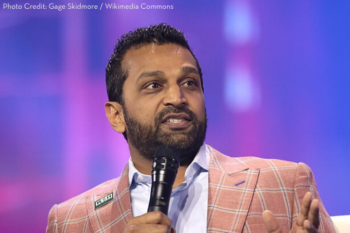 A man with a beard speaks into a microphone on stage, wearing a pink plaid blazer and a patterned shirt, with a purple and blue blurred background.